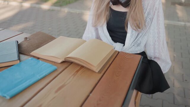 Close up of young woman sitting at outdoor wooden table with open notebooks and books. Woman interacts calmly turning pages in notebook while wearing headphones. scene features soft natural