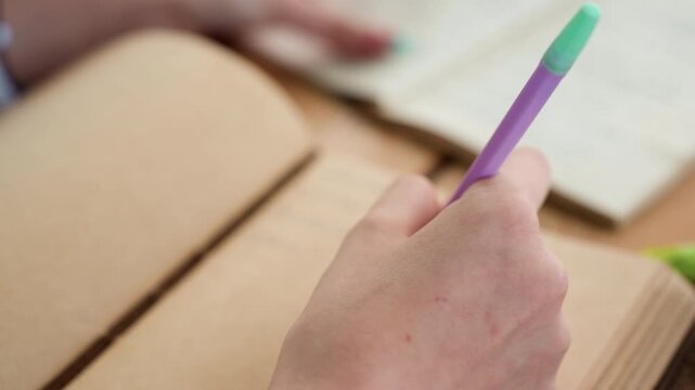 Close up of hand holding purple pen over open notebook with another person blurred hands visible in background. scene shows soft lighting and natural skin texture details focusing