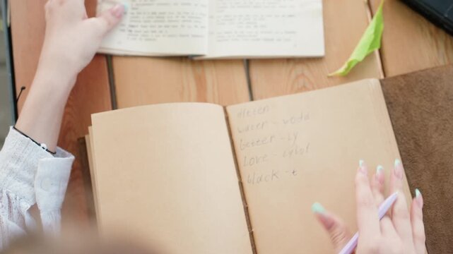 Close up of young woman writing in open notebook with handwritten words using pastel colored pen on wooden table. Background includes another book and green leaf detail soft natural lighting