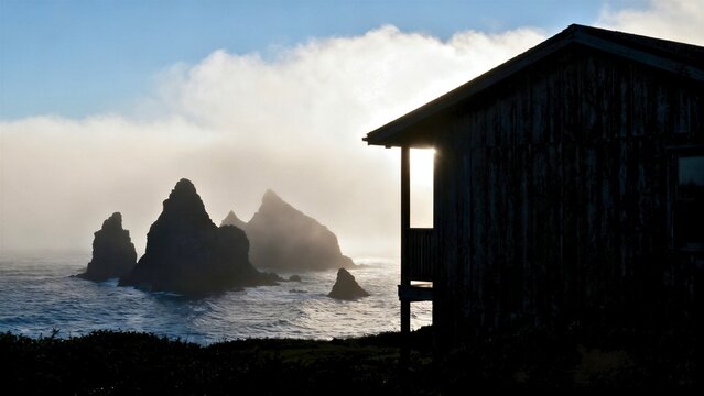 A dark charcoal-timber cabin sharply silhouetted against a bright, high-contrast fog bank, jagged black sea stacks piercing the mist offshore.