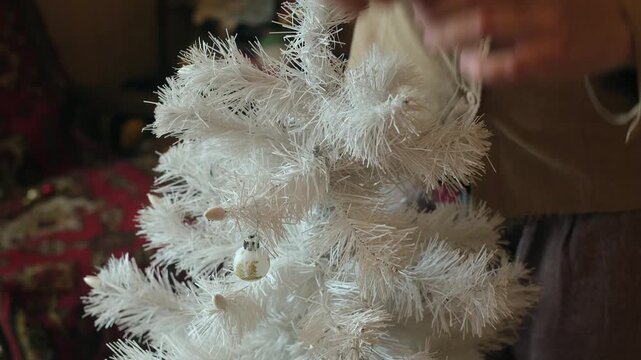 Female hands decorating white Christmas tree with Christmas decoration in cozy home.