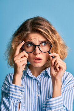 Junge Frau mit lockigem Haar schiebt runde Brille hoch, verwirrter Blick, blauer Hintergrund, ideal f&uuml;r Optik- und Sehtest-Werbung.