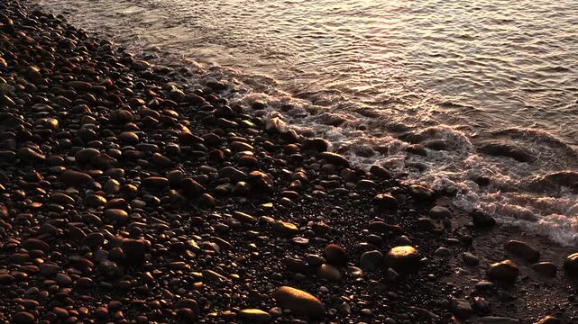 Golden sunset light reflecting on a rocky volcanic pebble beach with gentle ocean waves in Madeira, creating a peaceful and calming atmospheric coastal background. 4K video.