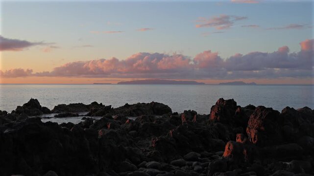Scenic view of rocky volcanic coastline in Madeira at sunset with dramatic golden twilight illuminating pink clouds over calm Atlantic ocean horizon. 4K video.