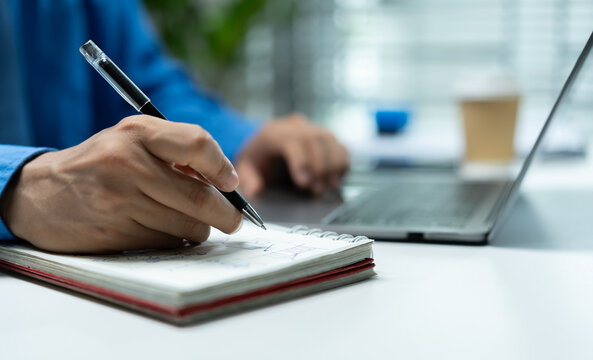 Businessman writing notes in a notebook beside a laptop, representing business planning, productivity, project management, brainstorming, organization and focused office work.
