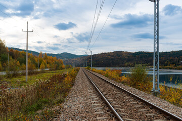 Straight electrified railway track follows the shore of Jezioro Mucharskie with autumn vegetation and forested Beskid hills in the distance. Commercial transportation scene combining rail infrastructu © Volha