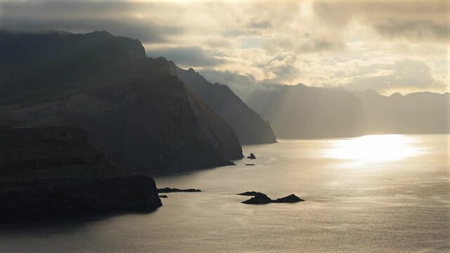 Dramatic golden sunbeams breaking through moody clouds over scenic volcanic ocean cliffs along Madeira coastline, creating a peaceful epic travel backdrop. 4K video.