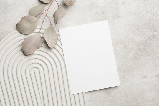 Blank white paper sheet positioned on a textured gray surface with a decorative curved mat and dried eucalyptus leaves for minimalist design and stationery mockup