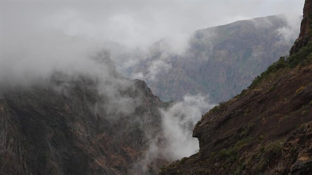 Dramatic moody fog swirling through rugged volcanic mountain peaks and deep steep valleys in Madeira island, creating an epic mystical wilderness landscape. 4K video.
