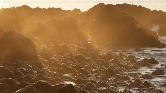 Golden sunset light shining through ocean mist over the rocky volcanic pebble coast with crashing waves in Madeira island, creating an ethereal peaceful mood. 4K video.