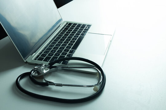 Stethoscope resting in front of an open laptop on a white desk with large copy space. Concept of telemedicine, online medical consultation, health tech, and computer repair service.