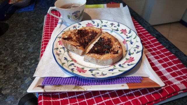 Close-up of a person having breakfast with toast covered in vegemite spread on a floral plate, accompanied by a cup of tea