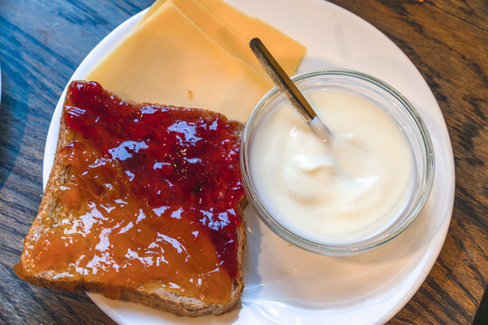 Toast bread spread with two fruit jams cheese slices and yogurt bowl on plate A white breakfast plate with a toasted slice of whole grain bread spread with red strawberry jam and orange marmalade