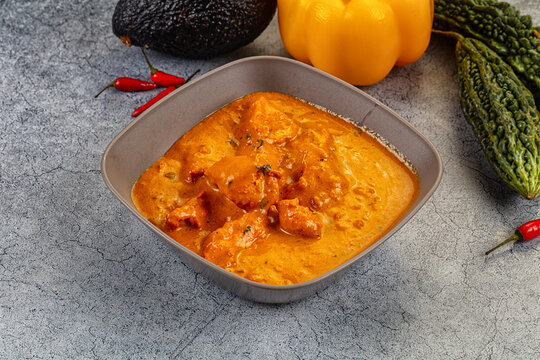 Creamy orange curry with chicken in square bowl surrounded by avocado yellow pepper and bitter gourd on textured gray surface