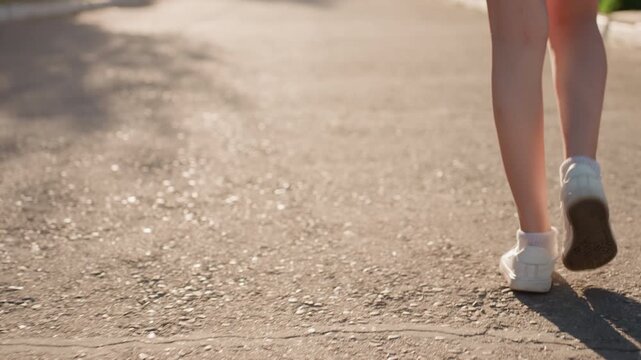 Close up of person legs and feet walking on outdoor path with white sneakers. focus remains steady capturing movement from heel to toe against sunlight dappled ground texture. Background