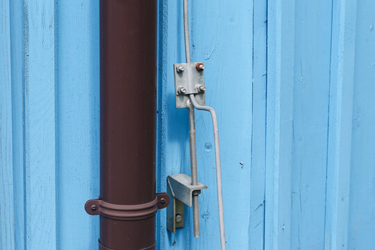 Metal lightning protection conductor and mounting hardware beside brown rainwater downpipe on blue wooden wall. Building electrical grounding detail and exterior construction background.