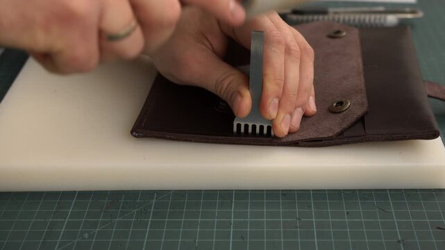 Leatherworker punching stitching holes in a brown leather wallet using a chisel and mallet in a workshop