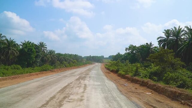 Reverse angle shot of rural dirt road under construction passing through palm oil sawit plantation and lush green tropical forest daylight transport infrastructure development