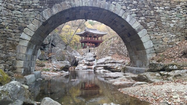 A scenic view of Seonseongyo, a designated treasure rainbow bridge at Suncheon Seonamsa Temple, with a traditional pavilion behind it. Clear valley water, colorful autumn foliage, and natural stones c