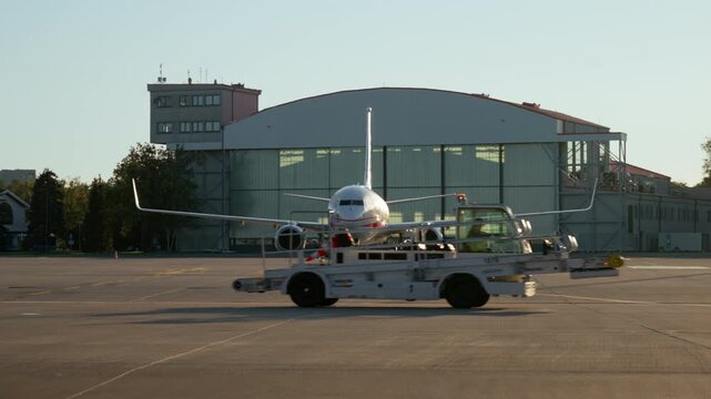 Passenger airplane stands on airport tarmac in front of a hangar. Ground support vehicle drives across the apron. Modern commercial airliner is ready for flight maintenance and air travel.