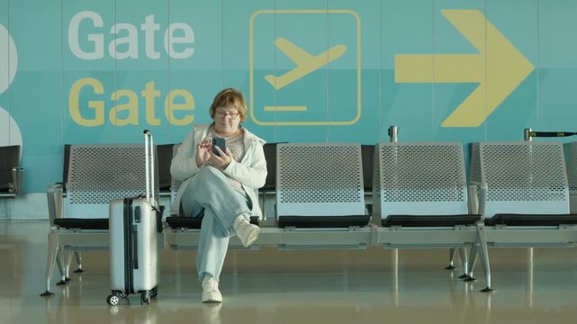 Mature woman sits with trolley suitcase at airport departure gate terminal. Elderly female passenger waits for delayed flight using phone. Modern transit hub provides quiet waiting lounge.