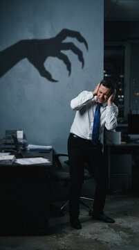 Scared businessman covering ears from a monstrous shadow, symbolizing fear or stress in a dark office environment, showcasing anxiety.