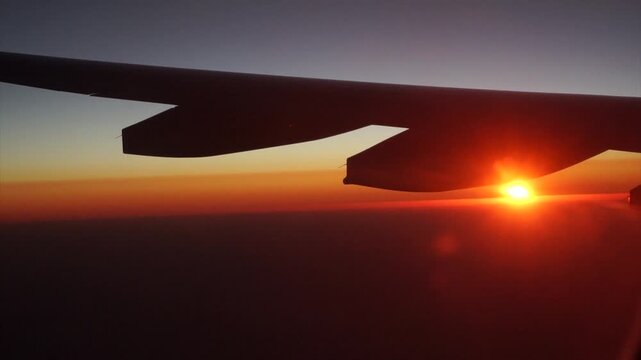 Close up shot of jet aircraft wing and engine silhouette hovering above clouds with brilliant golden sunburst at sunrise horizon through plane window for international airline travel footage