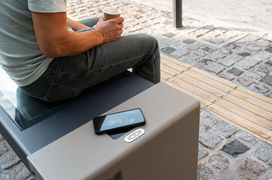 Man relaxes with coffee on smart solar-powered bench while his smartphone recharges wirelessly, illustrating modern lifestyle where both person and device recharge in sustainable urban infrastructure