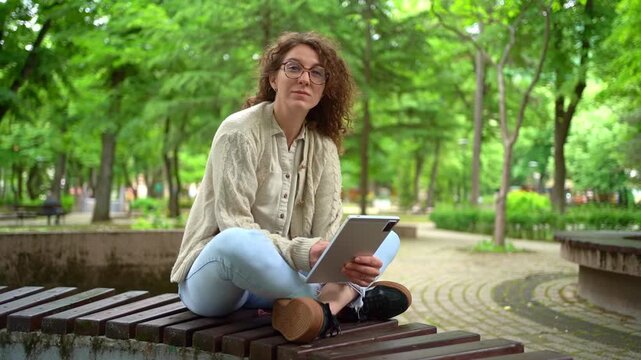 Woman with curly hair using tablet on a park bench
