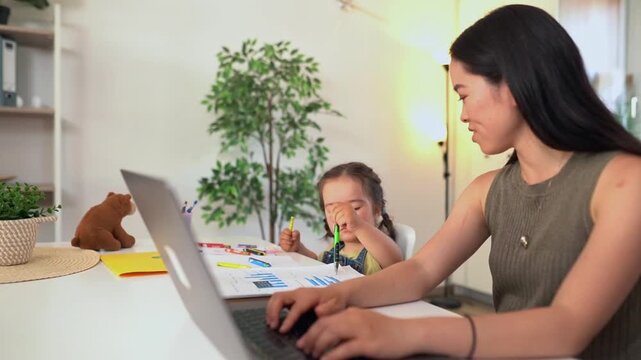 Japanese mother working on laptop with daughter drawing beside her
