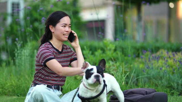 Japanese woman petting her french bulldog while talking on the phone in a park