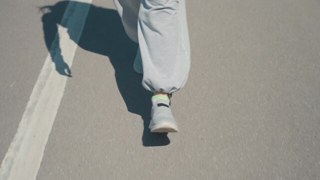 Close up of person legs and feet walking on outdoor paved surface. subject wears light gray sweatpants white slip on sneakers with green socks visible at ankle casting shadows under bright