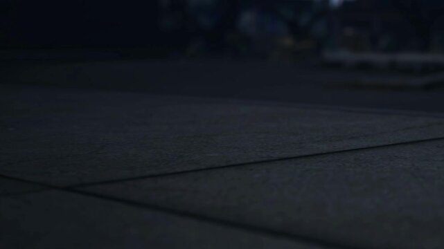 Footsteps echo on the concrete as a person walks alone at night. The surroundings are dimly lit, and the street appears empty and quiet.
