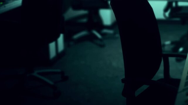 An office space is shown with empty chairs and computers turned off. The lights are dim and shadows fill the room, suggesting it is late at night.