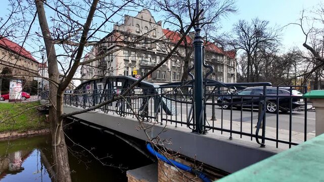 Video of a steel city bridge with decorative black railings over a river, showing historic tenement buildings, passing cars, and tree branches in the background on a sunny day.
