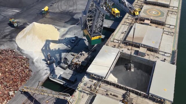 Aerial footage of a bulk cargo ship being loaded with dry material at an industrial port, showing a large crane, conveyor systems, cargo holds and dockside operations.