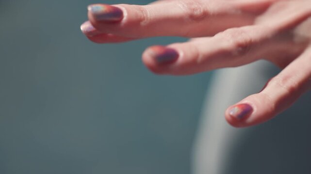 Close up of hand with iridescent nail polish moving gently against soft blue background. Hand remains in focus while fingers slightly shift position showcasing smooth skin texture and subtle