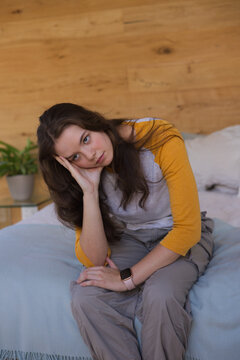 Adult woman sitting on bed leaning forward propping head, wearing mustard raglan tee and smartwatch