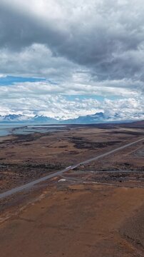 Aerial view of a scenic road stretching through the vast arid landscape of Patagonia, Argentina, with distant snow-capped mountains and a lake under a cloudy sky