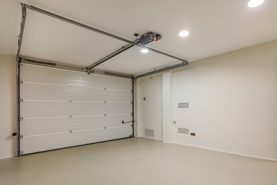 Clean empty residential garage with closed white sectional door bright overhead lighting and smooth light-colored floor