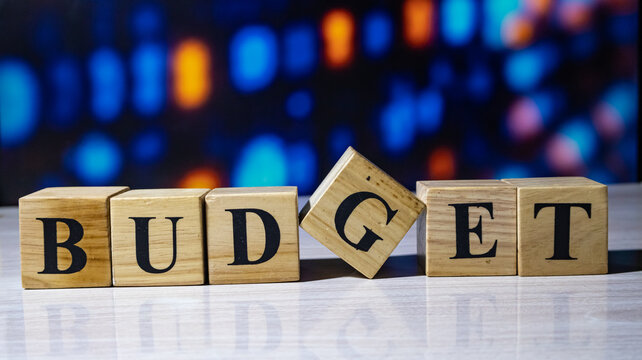 Wooden letter blocks spelling budget with a crooked block on glossy desk and bokeh background.