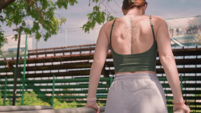 Woman with tattoo performs outdoor exercise routine on parallel bars in park setting wearing green sports bra and gray shorts. Background features metal bleachers trees fence clear sky