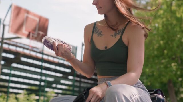 Close up of woman sitting on outdoor bench opening and examining contents of black drawstring bag. Woman is wearing casual athletic clothing with visible tattoos surrounded by greenery