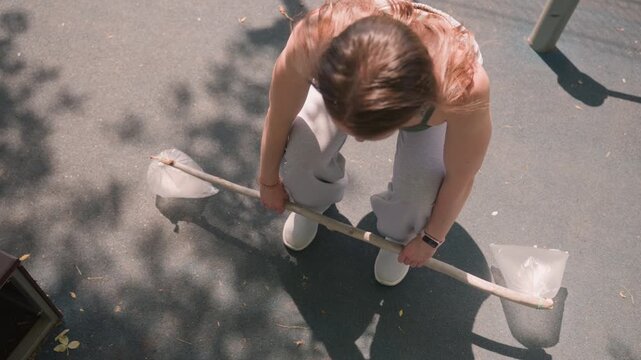 Close up of young woman holding and adjusting insect catching tool made from plastic bags sticks on paved ground with shadows. subject wears light gray sweatpants white shoes and has visible