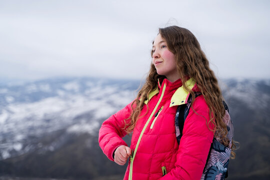 A woman in a warm pink winter jacket is preparing for a mountain hike, surrounded by snow-covered peaks. Female traveler zips up her jacket. Winter trekking and outdoor adventure concept.