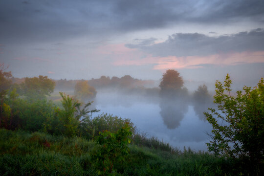 Morning fog drifting over a calm river surrounded by summer greenery