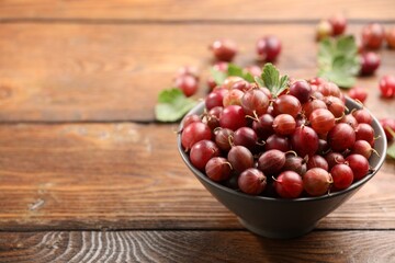 Fresh red gooseberries in bowl and green leaves on wooden table, closeup. Space for text © New Africa