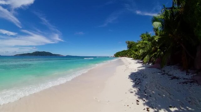 Expansive white sand beach stretching alongside turquoise ocean waves, with palm trees and distant islands. Seychelles, La Digue.