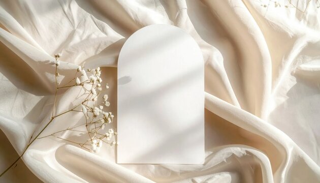 Arched paper card mockup with delicate baby breath flowers on draped beige fabric backdrop, wedding stationery invitation display