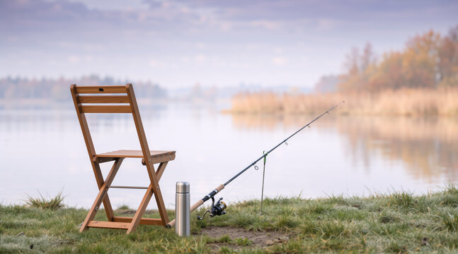 Wooden folding chair with a fishing rod and silver thermos on a grassy lake shore. Misty morning autumn fishing setup